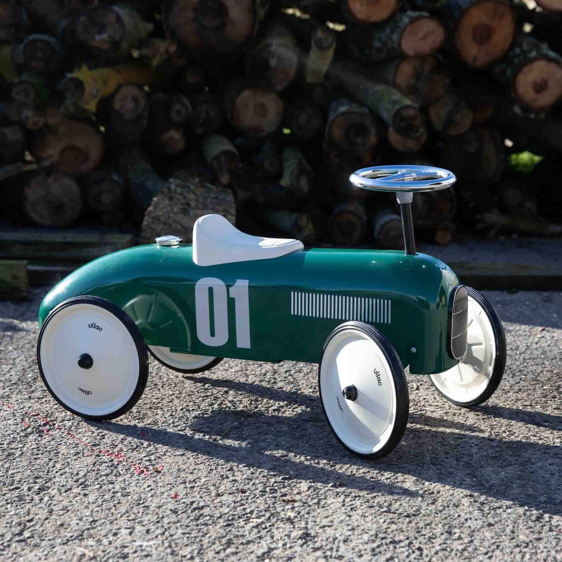 A green ride-on toy car placed on a gravel surface outdoors, with stacked logs in the background and sunlight casting shadows across the ground.