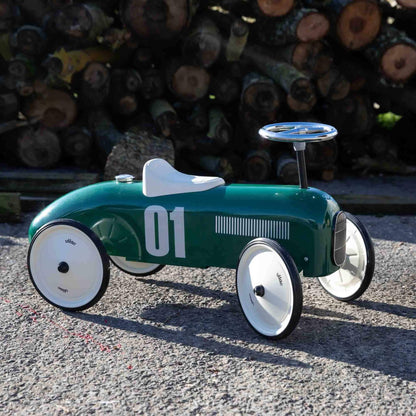 A green ride-on toy car placed on a gravel surface outdoors, with stacked logs in the background and sunlight casting shadows across the ground.