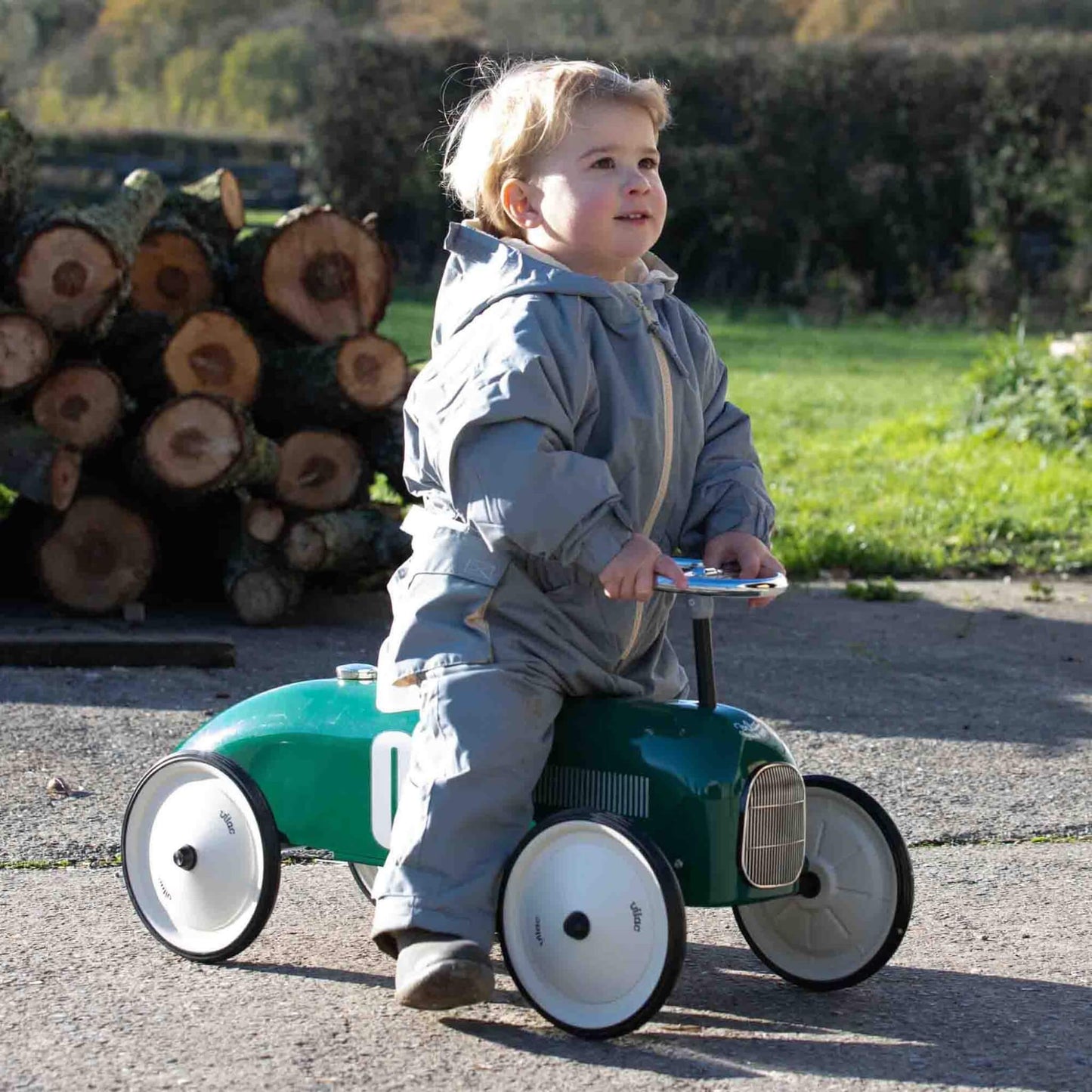 A young child sitting on a green ride-on toy car outdoors, wearing a padded grey outfit and holding the steering wheel while moving along a paved surface.
