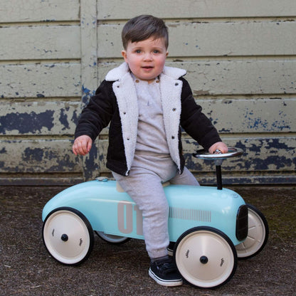 A young child wearing a grey outfit and black jacket with a white fleece collar is sitting on a pale blue metal ride-on toy car with cream wheels, holding the small steering wheel. The child is looking forward and smiling slightly, with a weathered wooden wall behind them.