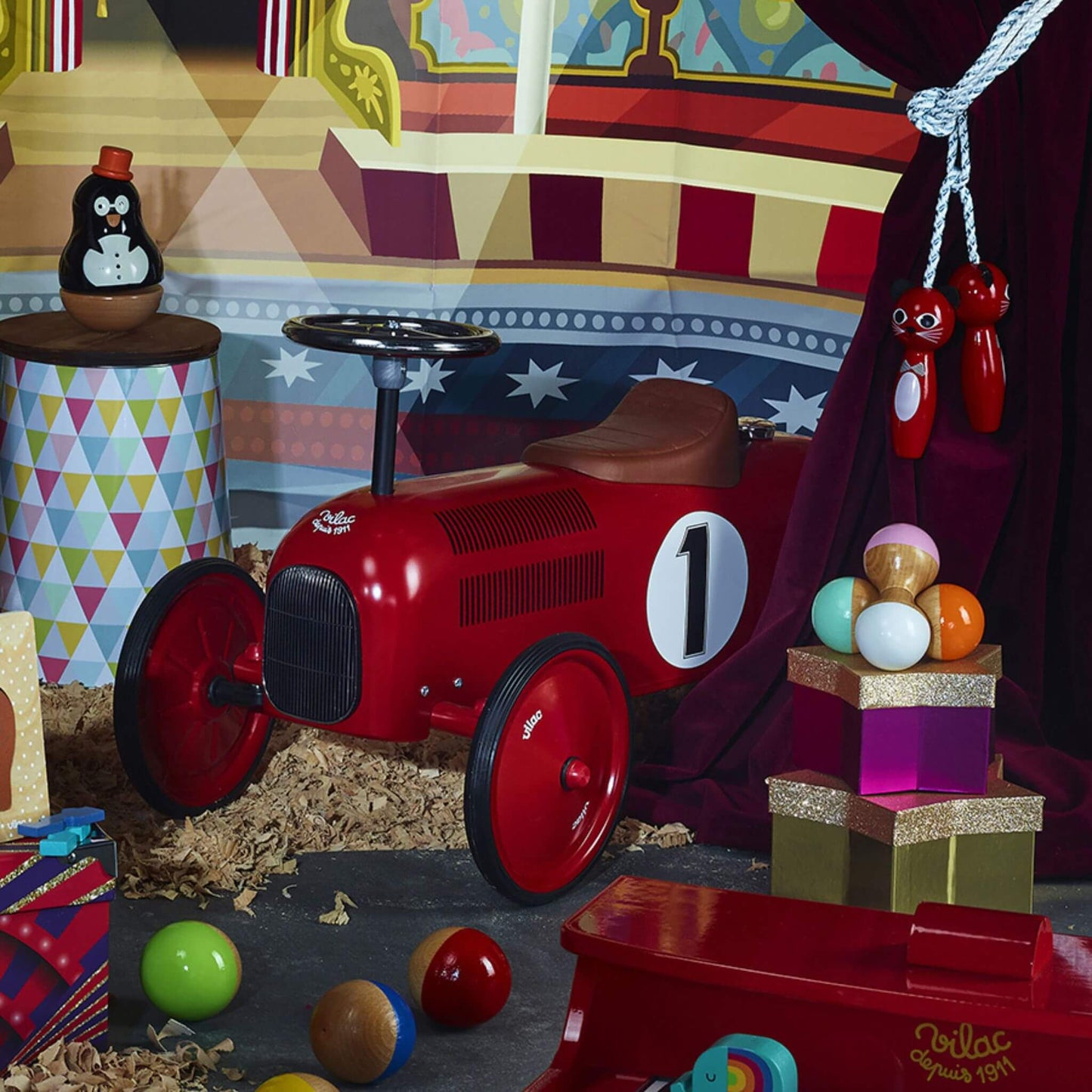 A red metal ride-on car placed in a colourful playroom with toys and circus-themed decorations around it.