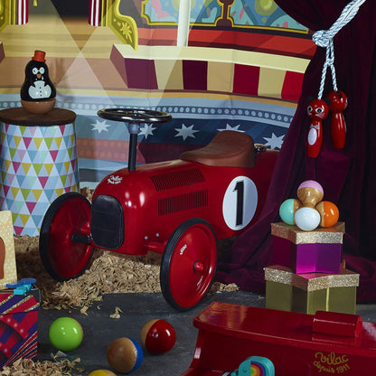 A red metal ride-on car placed in a colourful playroom with toys and circus-themed decorations around it.