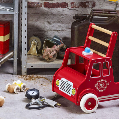 A wooden fire engine walker placed in a playroom setting with toys around it, showing real-life use as a toddler push along walker.