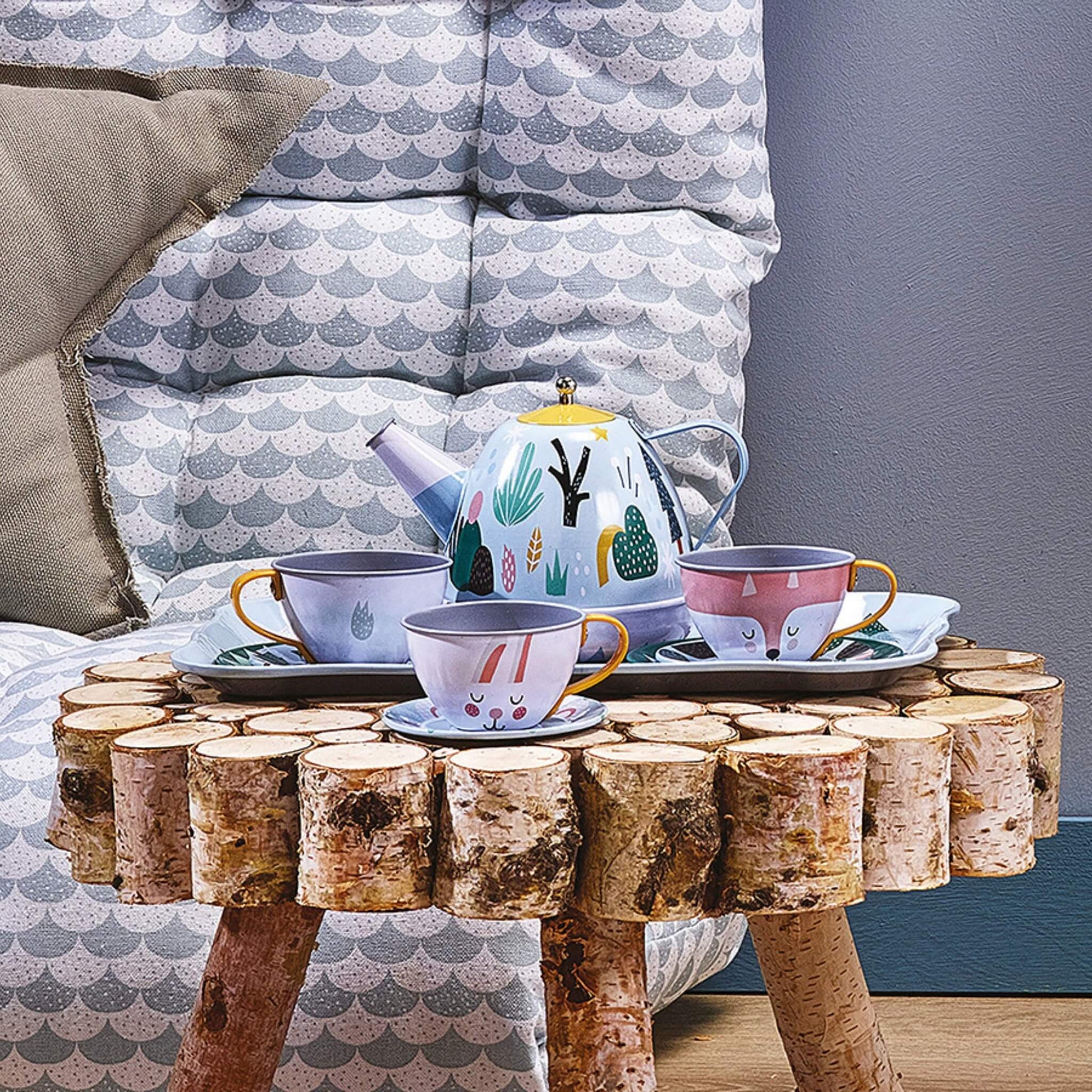 A children’s tin tea set with a decorated tray, teapot and four cups placed on a rustic wooden stool in a cosy room setting.