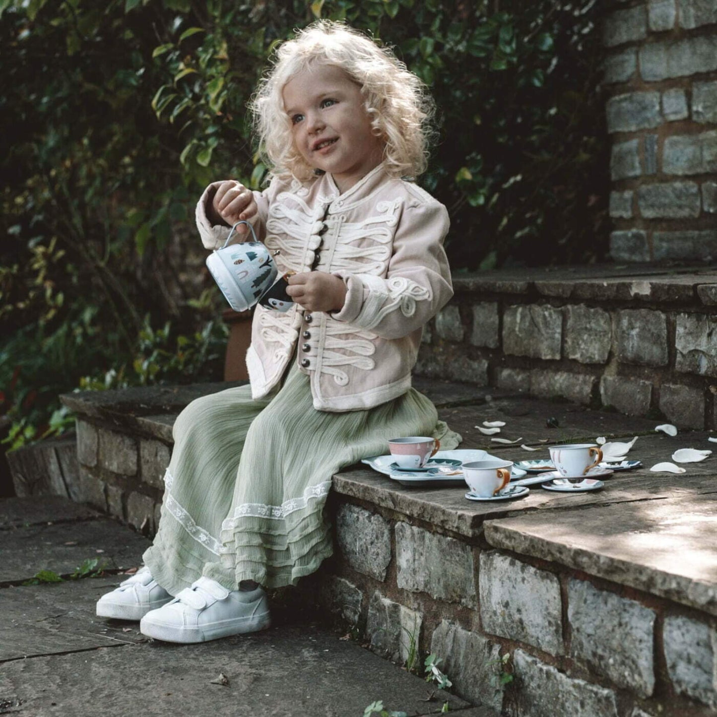 A young child sitting on stone steps holding a tin teapot while playing with matching cups and saucers arranged beside her.