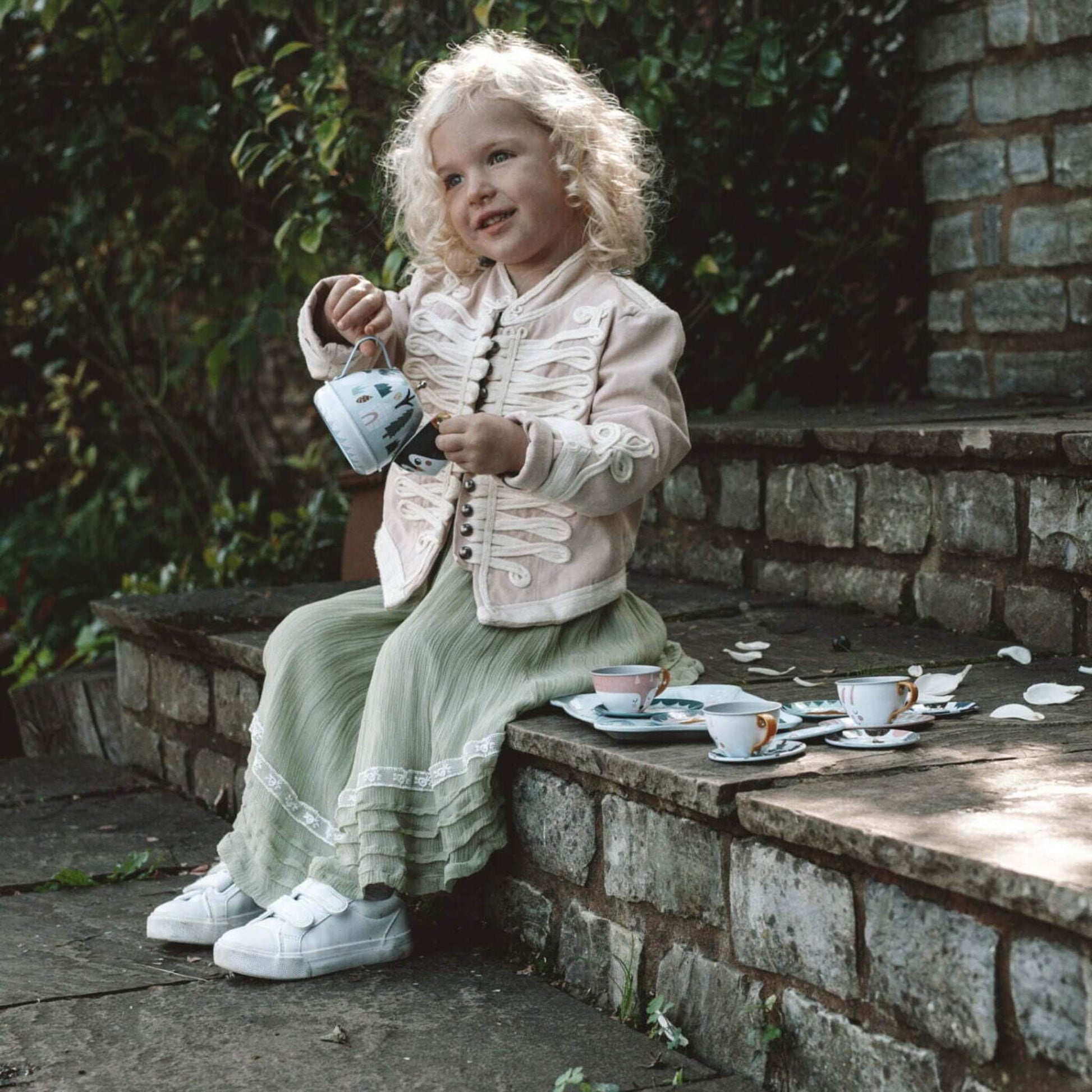 A young child sitting on stone steps holding a tin teapot while playing with matching cups and saucers arranged beside her.