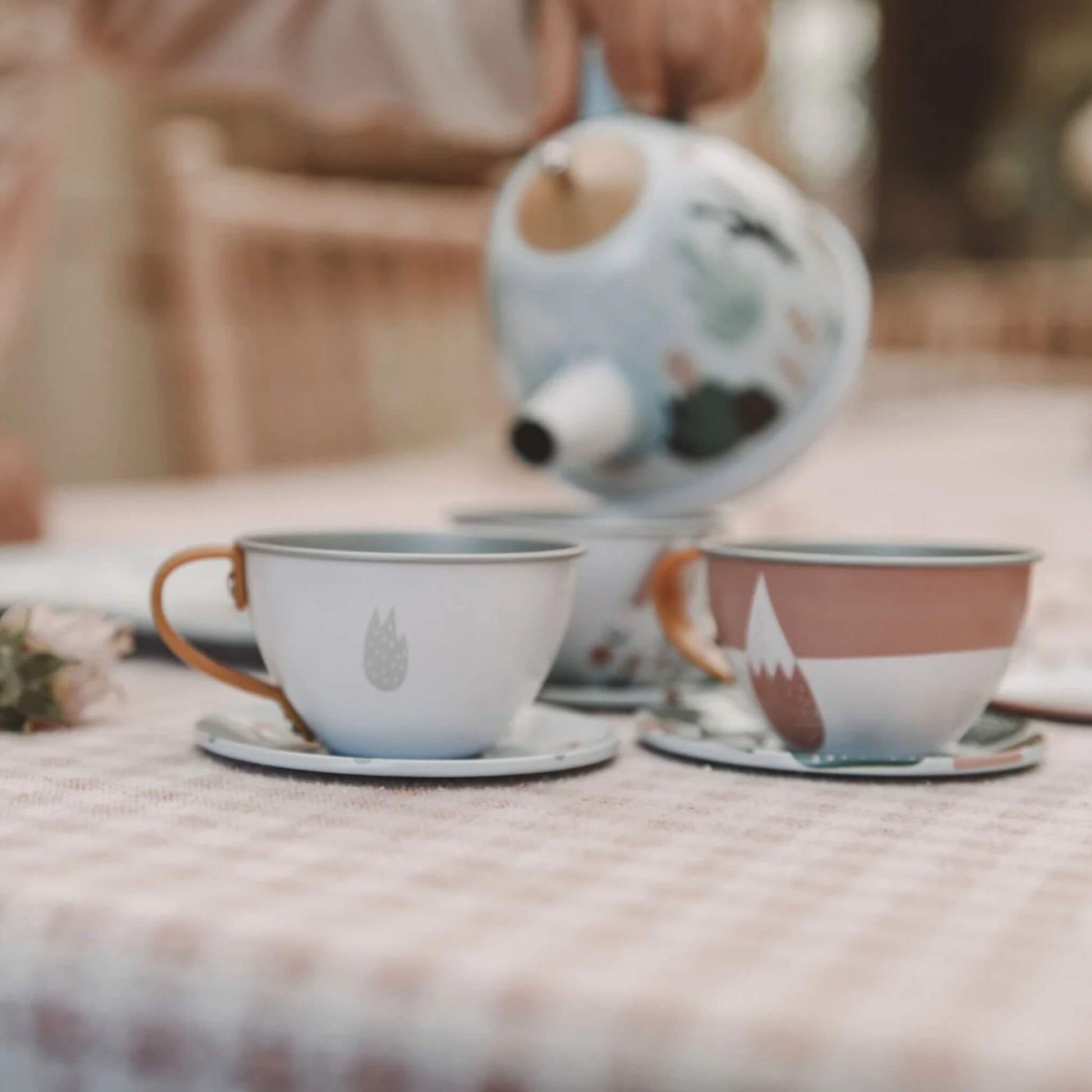 A close-up of tin teacups on saucers arranged on a checkered tablecloth while a hand pours from the decorated teapot.