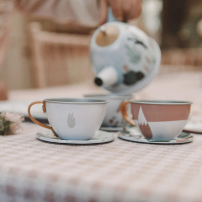 A close-up of tin teacups on saucers arranged on a checkered tablecloth while a hand pours from the decorated teapot.