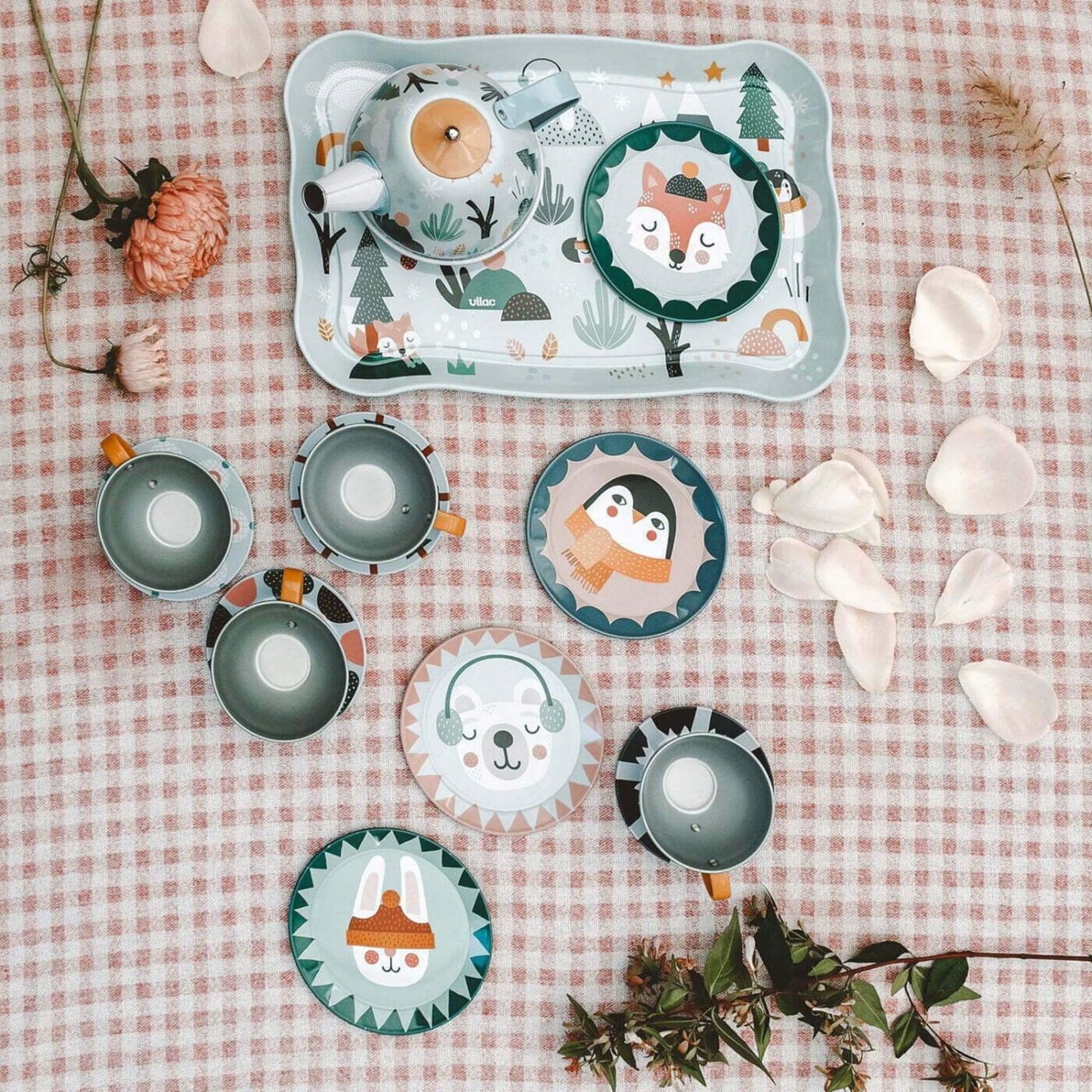 A full tin tea set laid out on a pink checkered tablecloth with a decorated tray, four cups and saucers, and illustrated plates featuring animals.