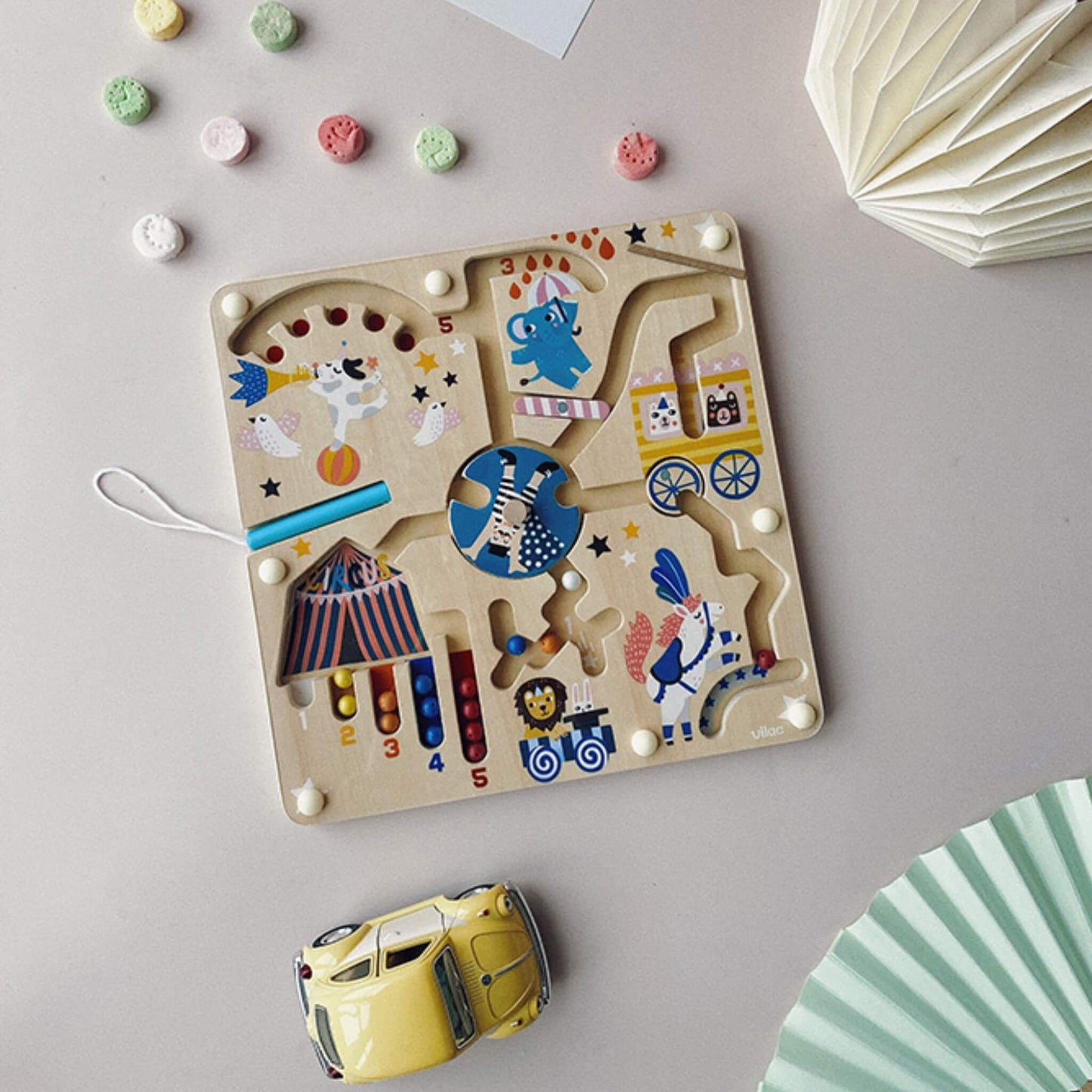 Circus-themed magnetic maze toy laid flat on a table with sweets and a yellow toy car nearby, showing the maze board in use with its stylus attached.
