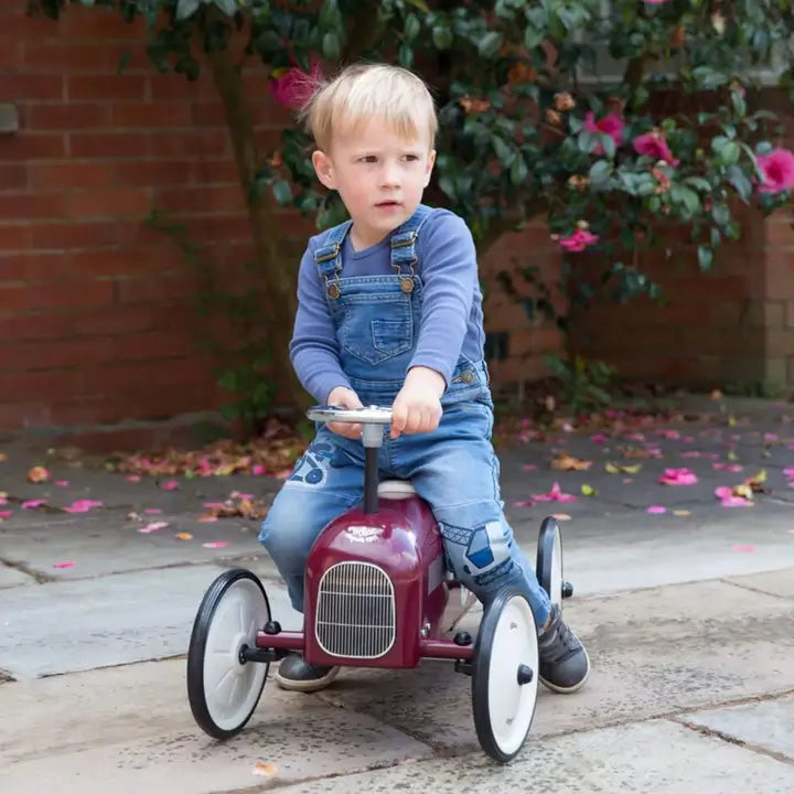A young boy in denim dungarees riding a burgundy metal ride-on car outdoors, holding the steering wheel while looking to the side.