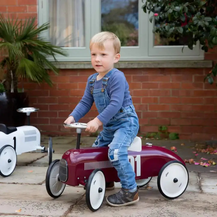 A young boy in denim dungarees riding a burgundy ride-on toy car outside, pushing forward with his feet while holding the steering wheel.