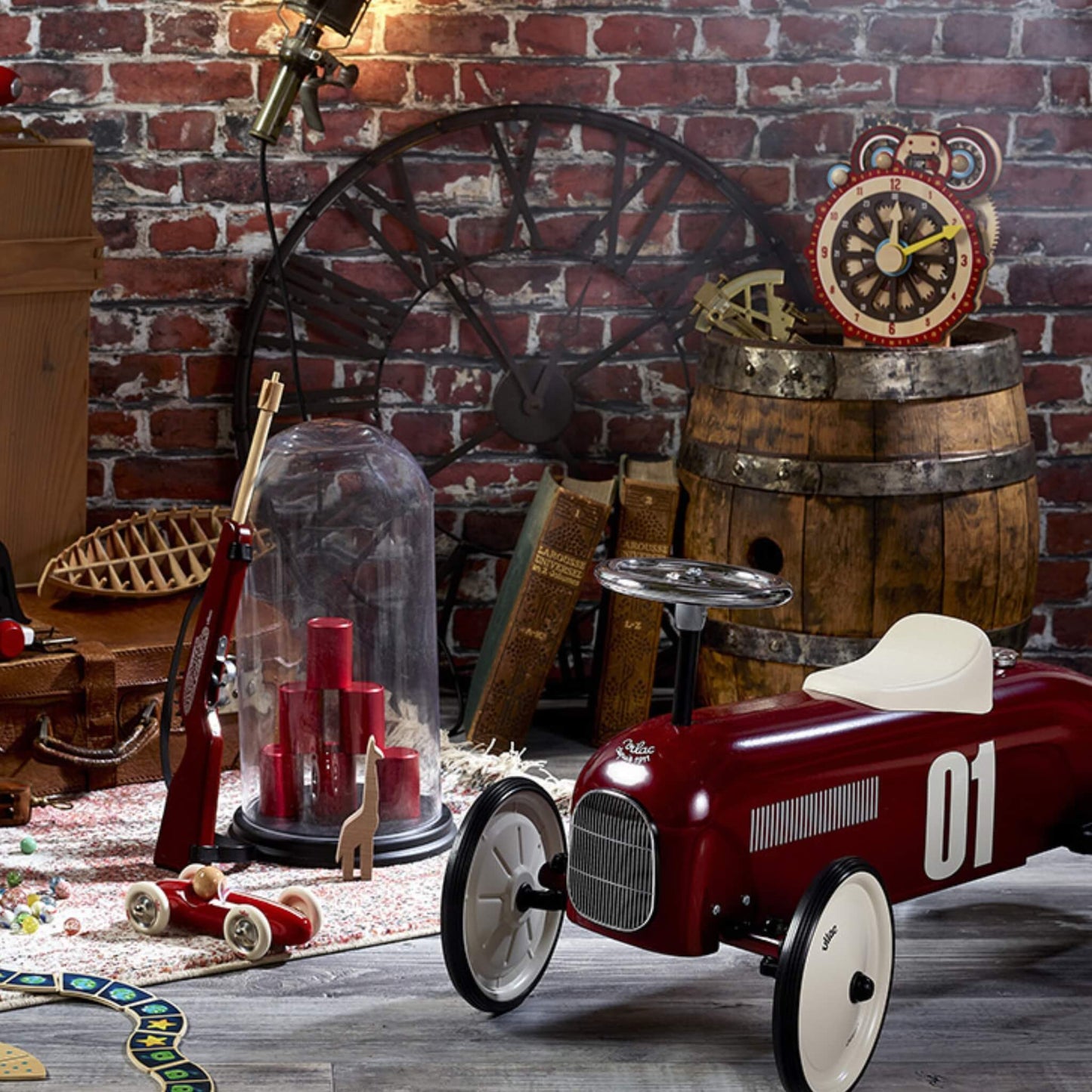 A burgundy ride-on toy car with number 01 decal displayed indoors among vintage-style décor including books, a wooden barrel and a wall clock.