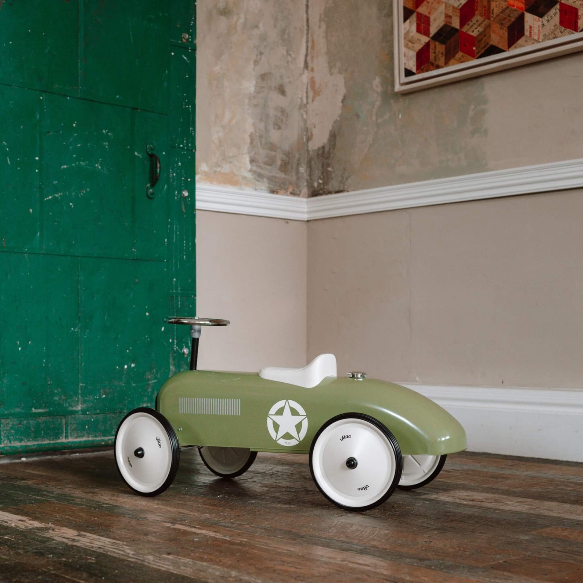 A green metal ride-on car with four cream wheels, white seat and steering wheel, placed on a wooden floor against a rustic wall.