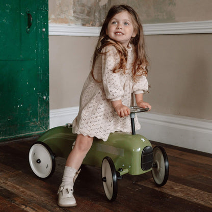 A young girl in a cream dress standing on a green metal ride-on toy car, holding the steering wheel with both hands.
