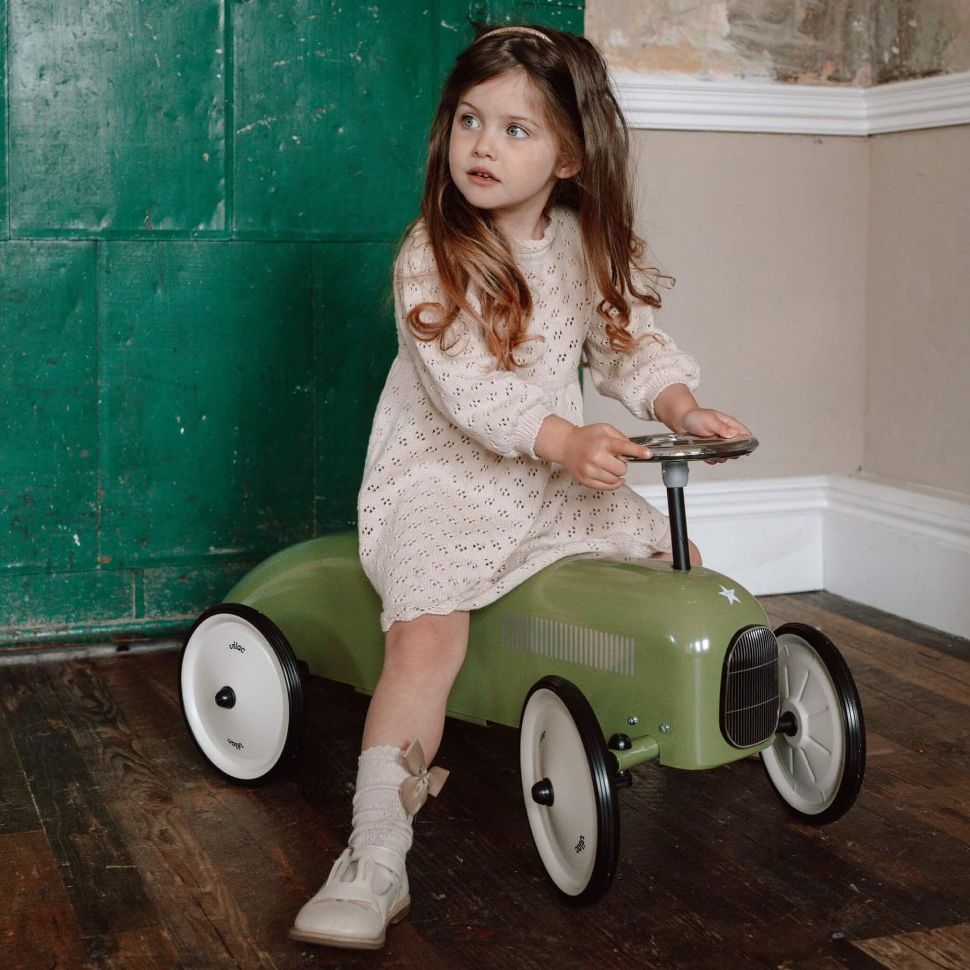 A young girl in a cream dress sitting on a green ride-on toy car, holding the steering wheel while looking to the side.