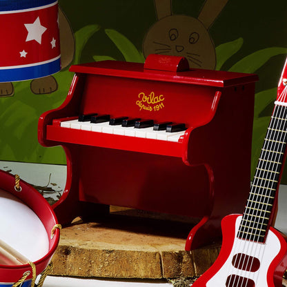 Red wooden toy piano with black and white keys placed in a playroom setting, shown alongside a toy drum and red guitar against a painted animal backdrop.