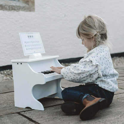 Young child sitting outdoors on a stone patio, playing a white toy piano with sheet music displayed above the keys.