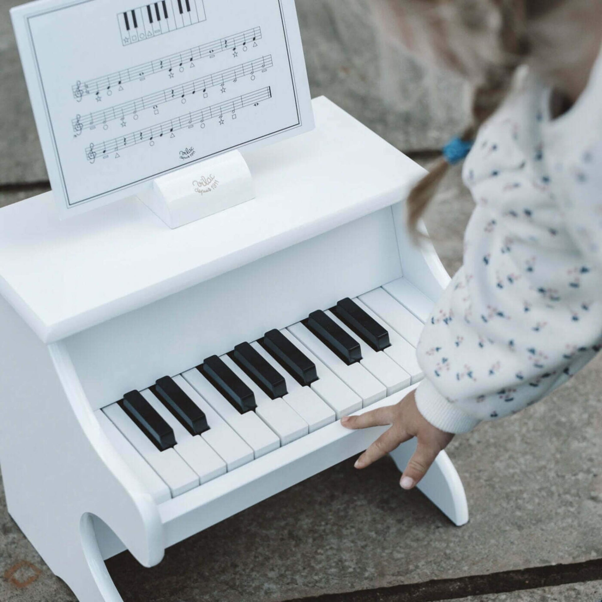 Child leaning forward to play a white wooden toy piano outdoors, with a music score card set in the holder above the keys.