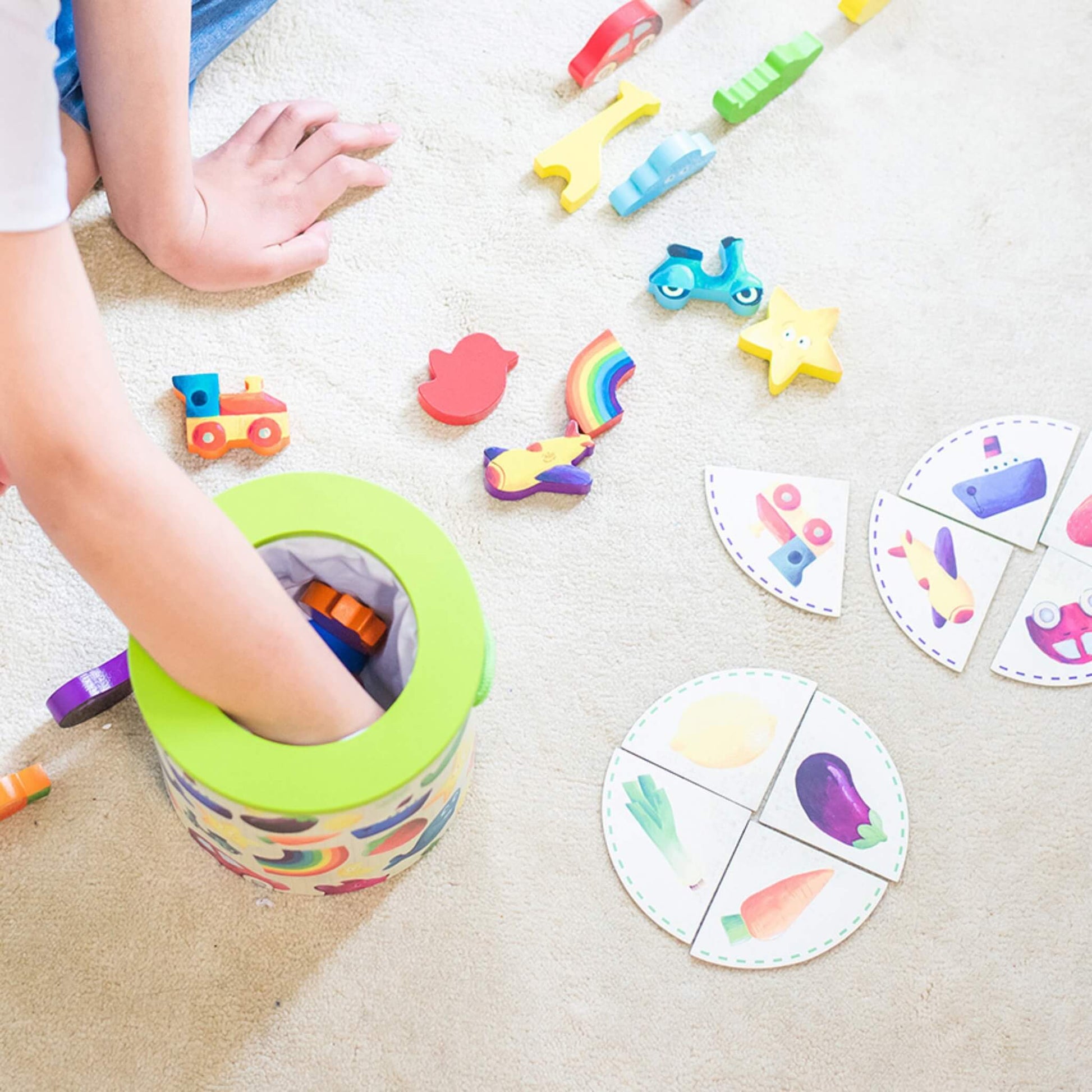 Child reaching into a toy barrel to pick out wooden pieces, with illustrated cards and colourful game pieces arranged on the floor.