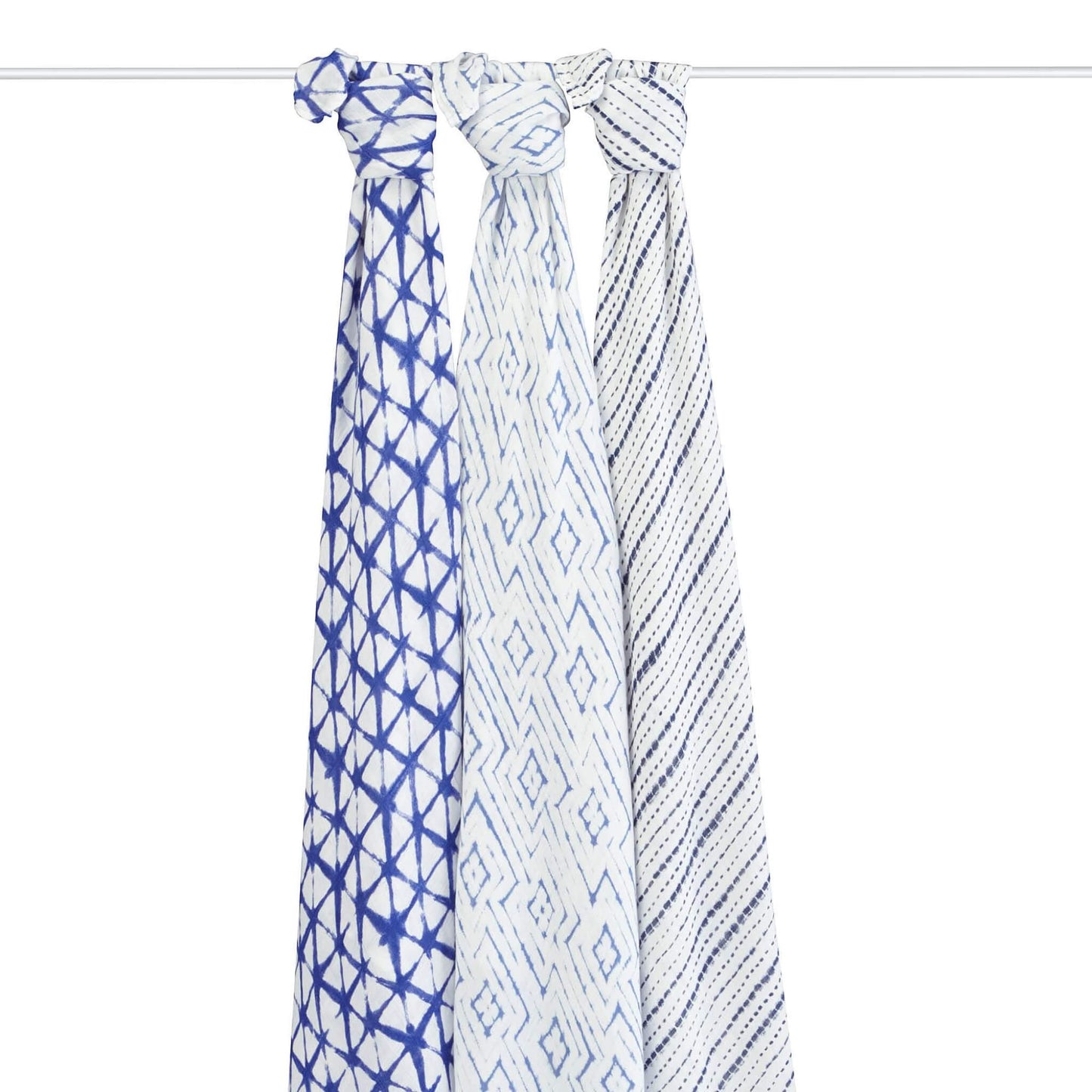 Three muslin swaddles hang knotted over a rod, each featuring a different indigo pattern: a starburst grid design, a diamond tile print, and a broken stripe pattern.