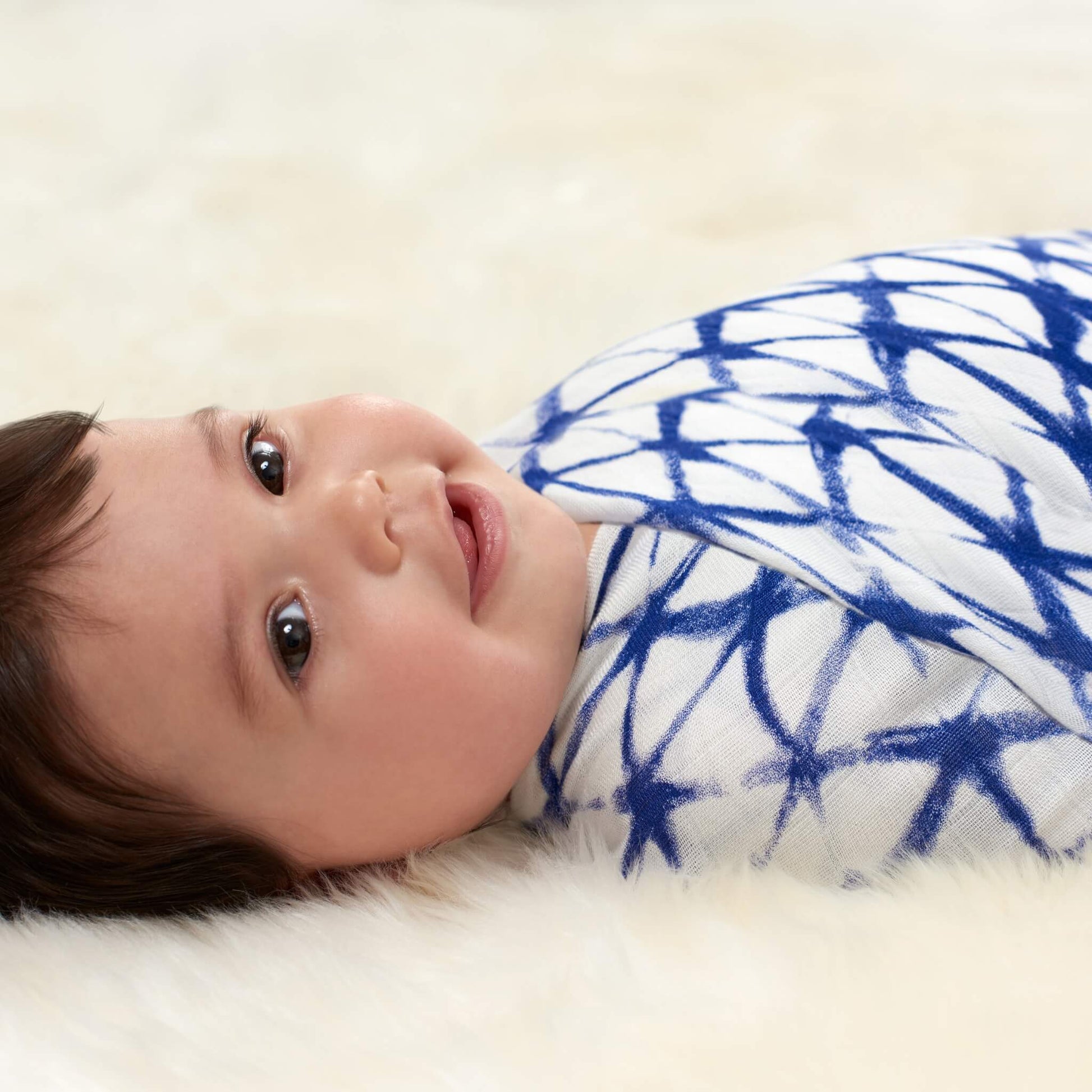 A baby lies on a soft cream surface swaddled in a white muslin blanket with a deep indigo geometric starburst pattern, facing upwards and smiling gently.