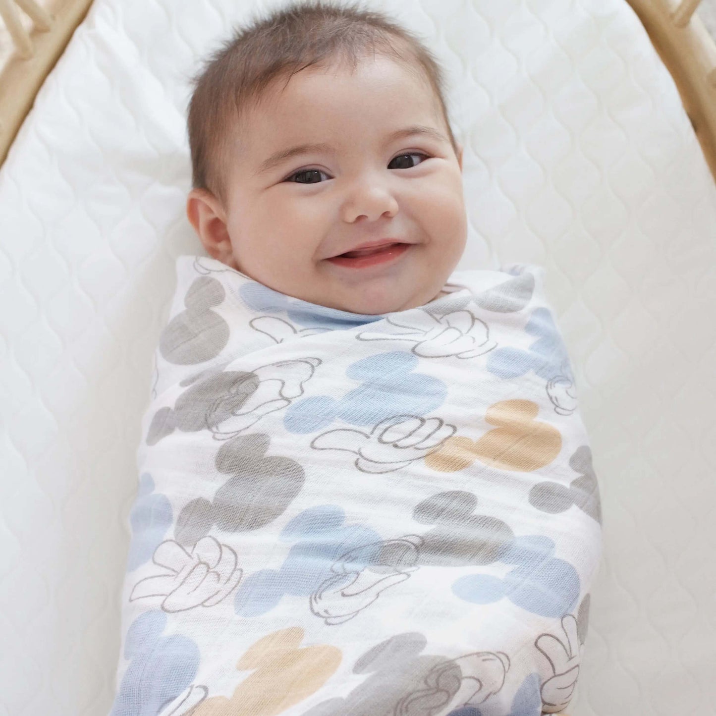 Smiling baby swaddled in aden + anais cotton muslin swaddle featuring Mickey Mouse print in soft blue and beige.