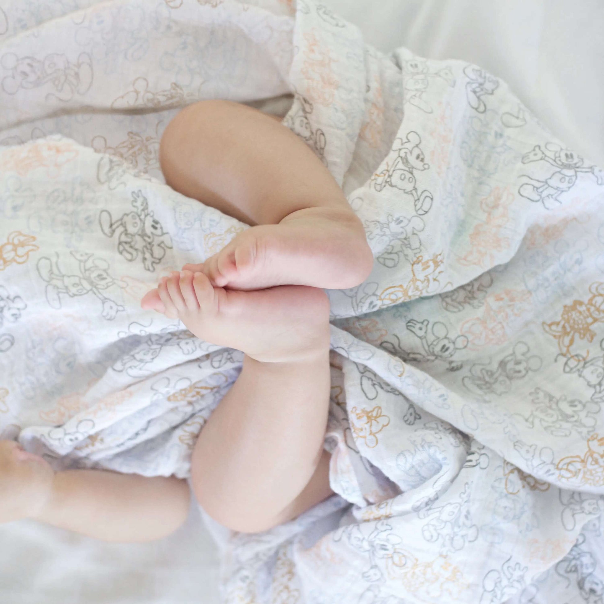 Close-up of baby’s feet wrapped in aden + anais muslin swaddle with delicate Minnie Mouse print in gold and grey.