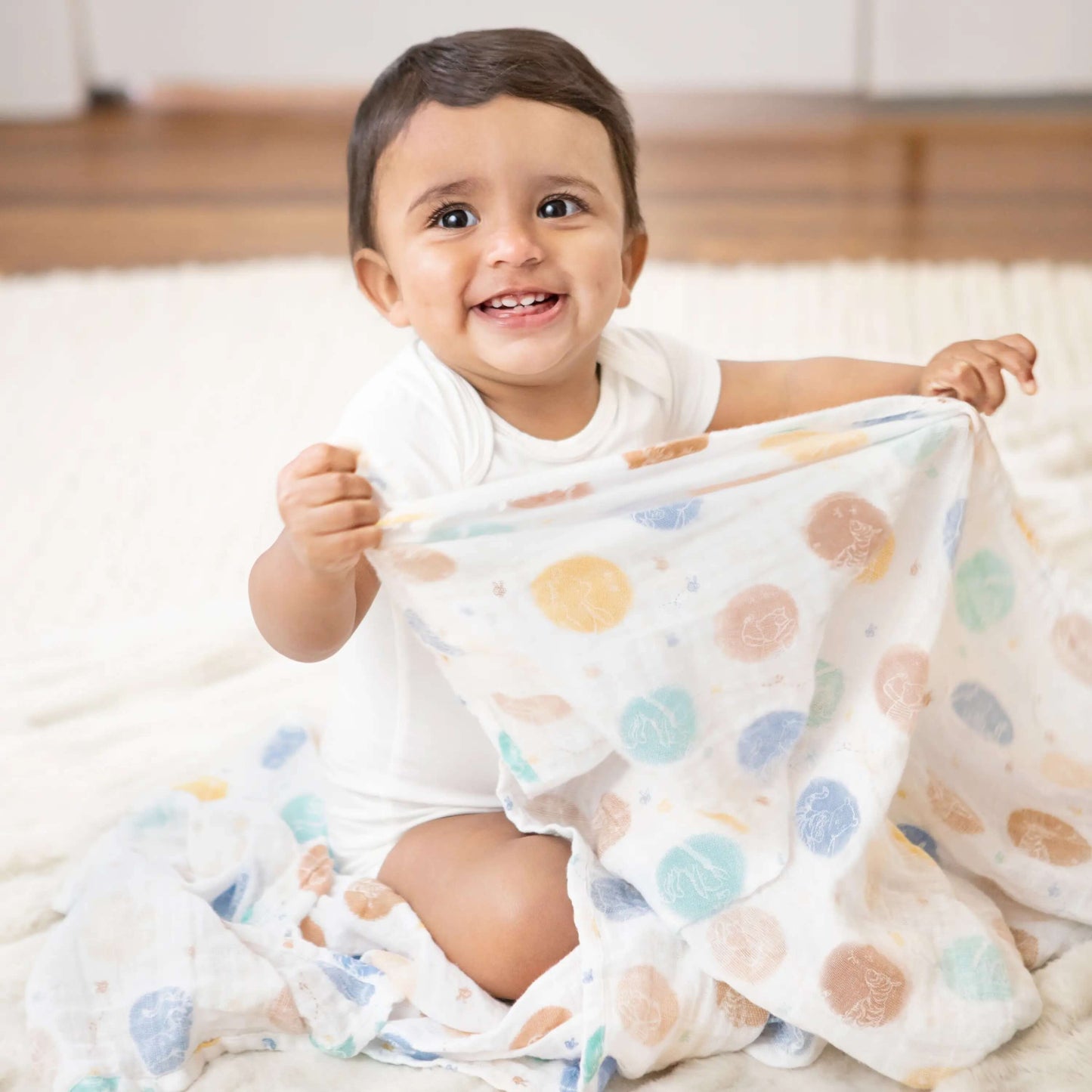 Baby sitting and holding aden + anais swaddle featuring Winnie the Pooh print in multicoloured dot pattern on soft white muslin.