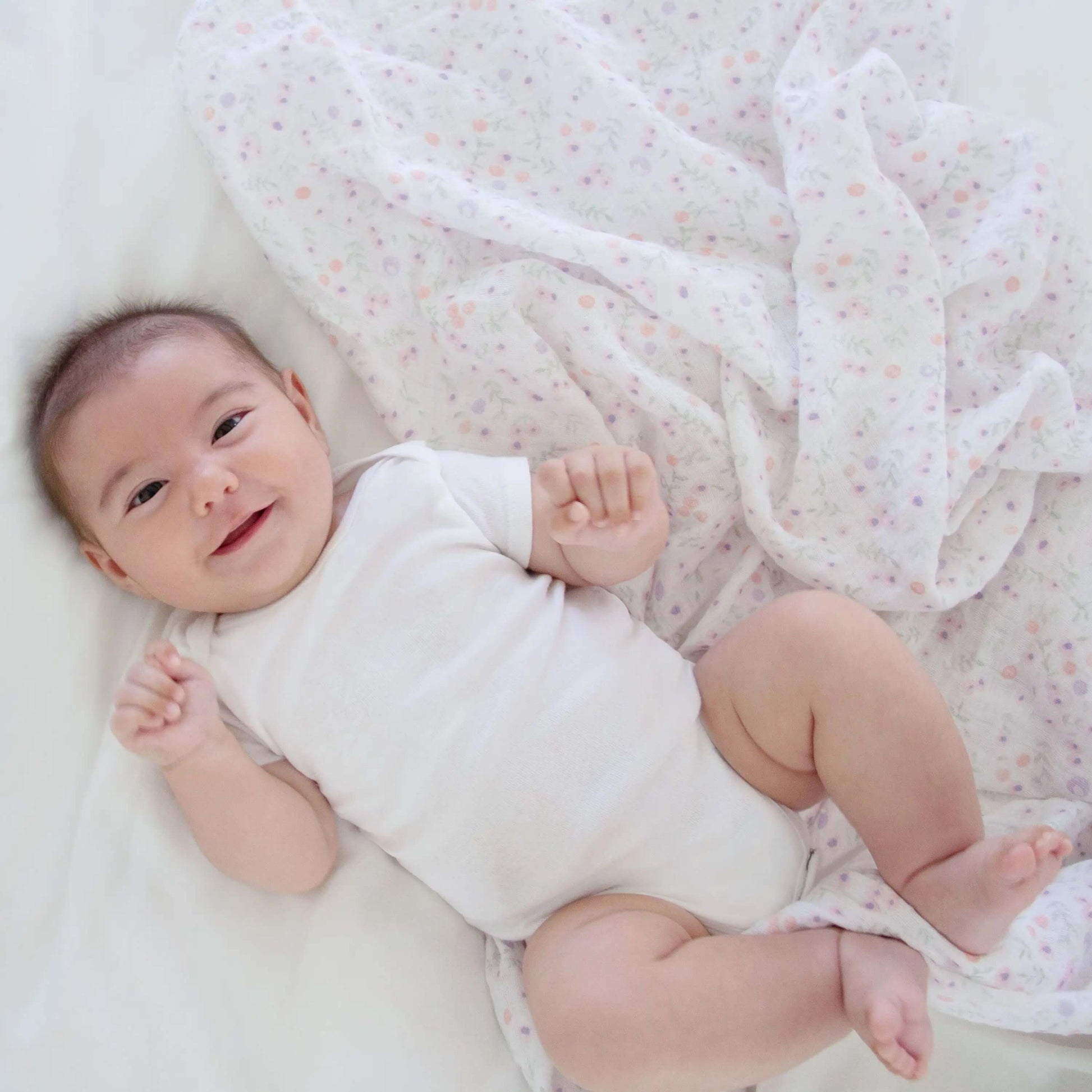 Smiling baby lying on bed with aden + anais Ma Fleur muslin swaddle with pastel dots.