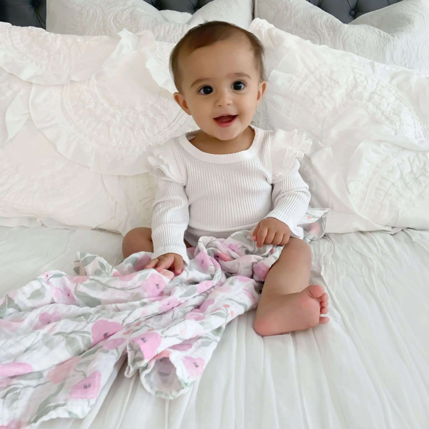 Baby sitting up on white bed holding aden + anais Ma Fleur swaddle in rose print.