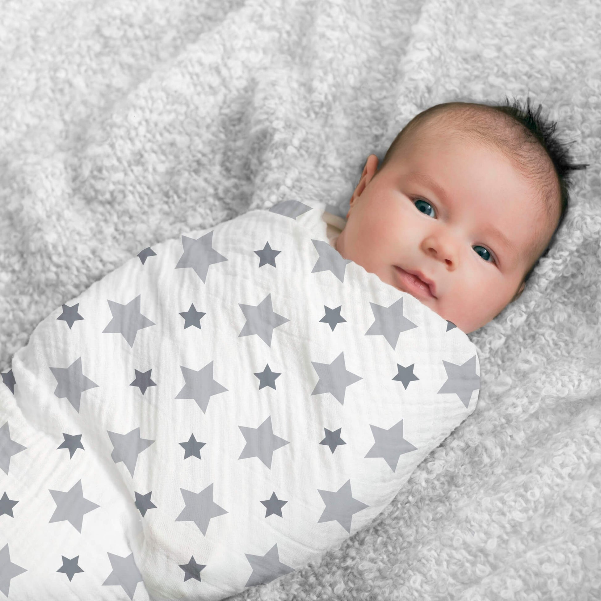 Baby awake and smiling, swaddled in grey star aden + anais cotton muslin. Perfect for soft, breathable newborn comfort.