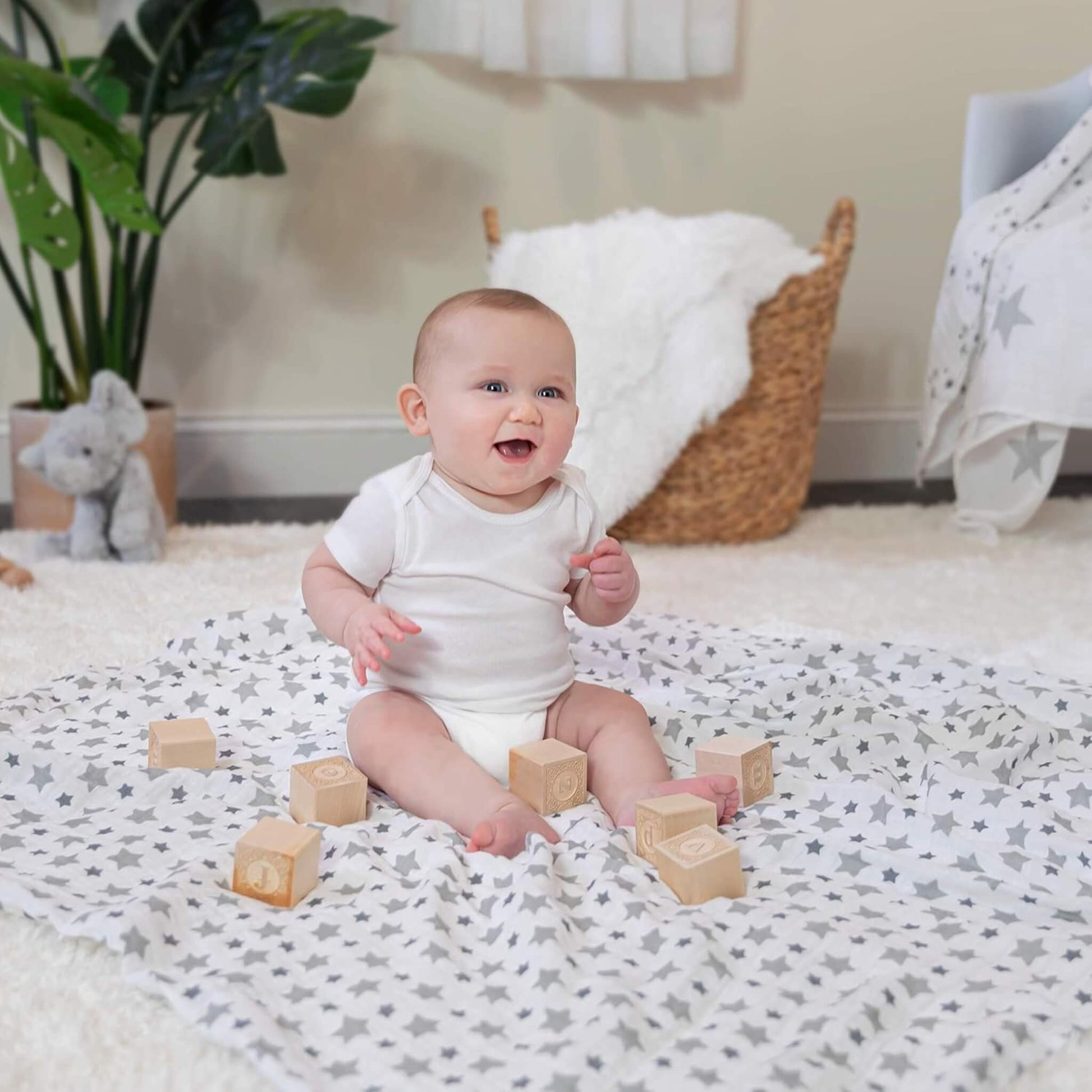 Smiling baby sitting on aden + anais Twinkle muslin swaddle. Surrounded by wooden blocks in a soft nursery setting. Lightweight and breathable cotton.