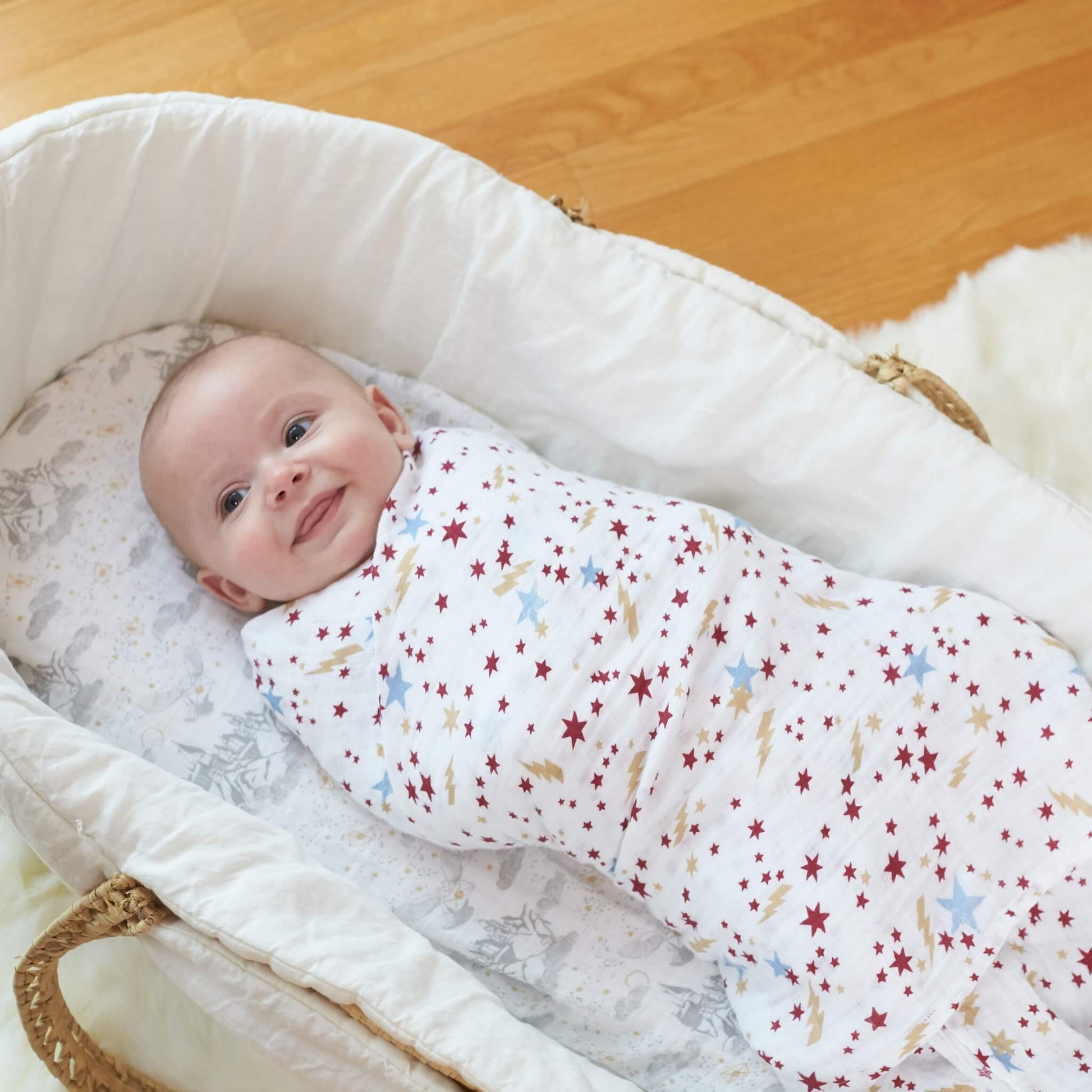 Smiling baby swaddled in the star print aden + anais Harry Potter™ muslin swaddle, lying in a Moses basket with themed bedding.