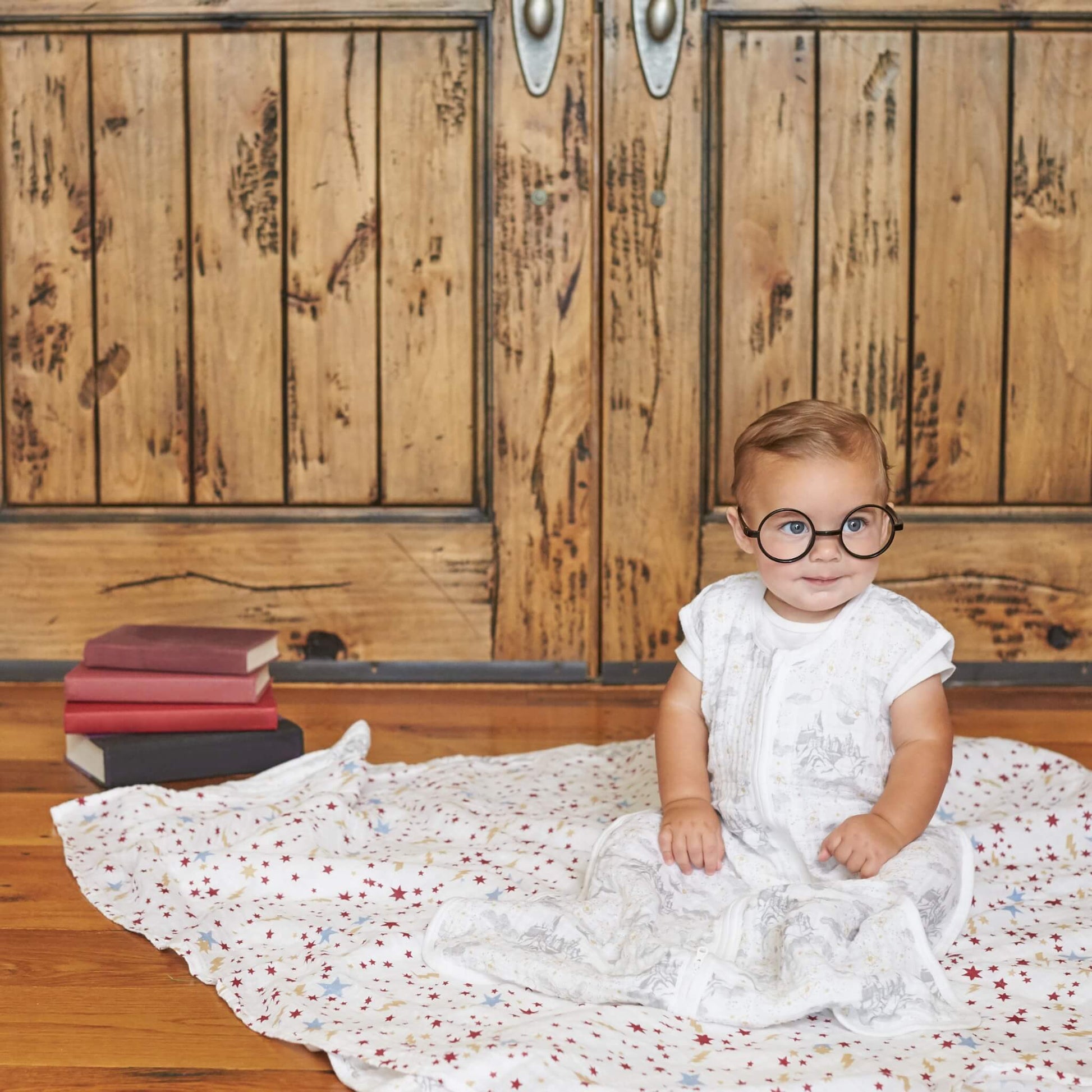 Toddler wearing round glasses sitting on an aden + anais Harry Potter™ swaddle used as a blanket, surrounded by books and wooden decor.