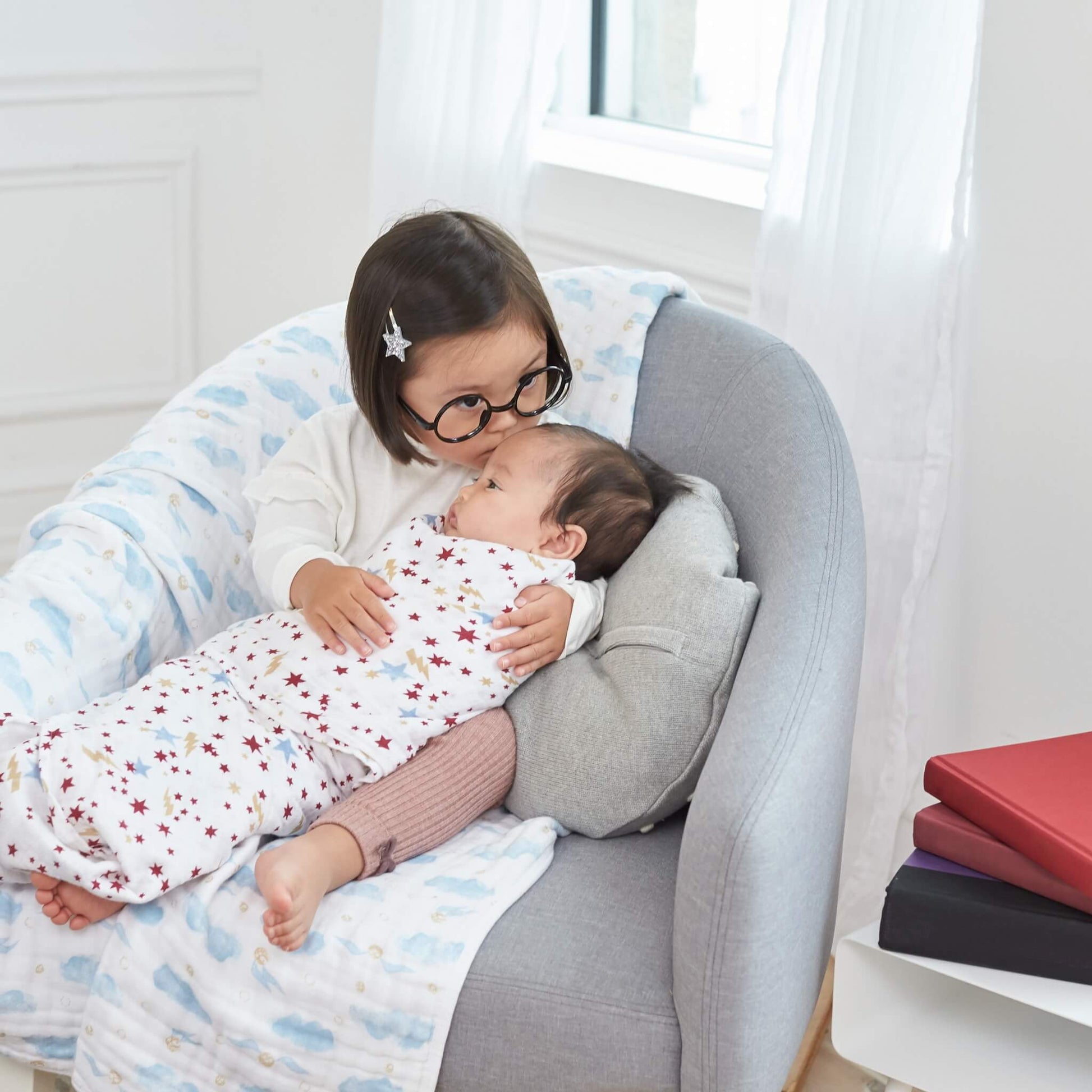 Older child cuddling baby swaddled in aden + anais Harry Potter™ star print muslin while seated on a grey chair with another swaddle draped over.
