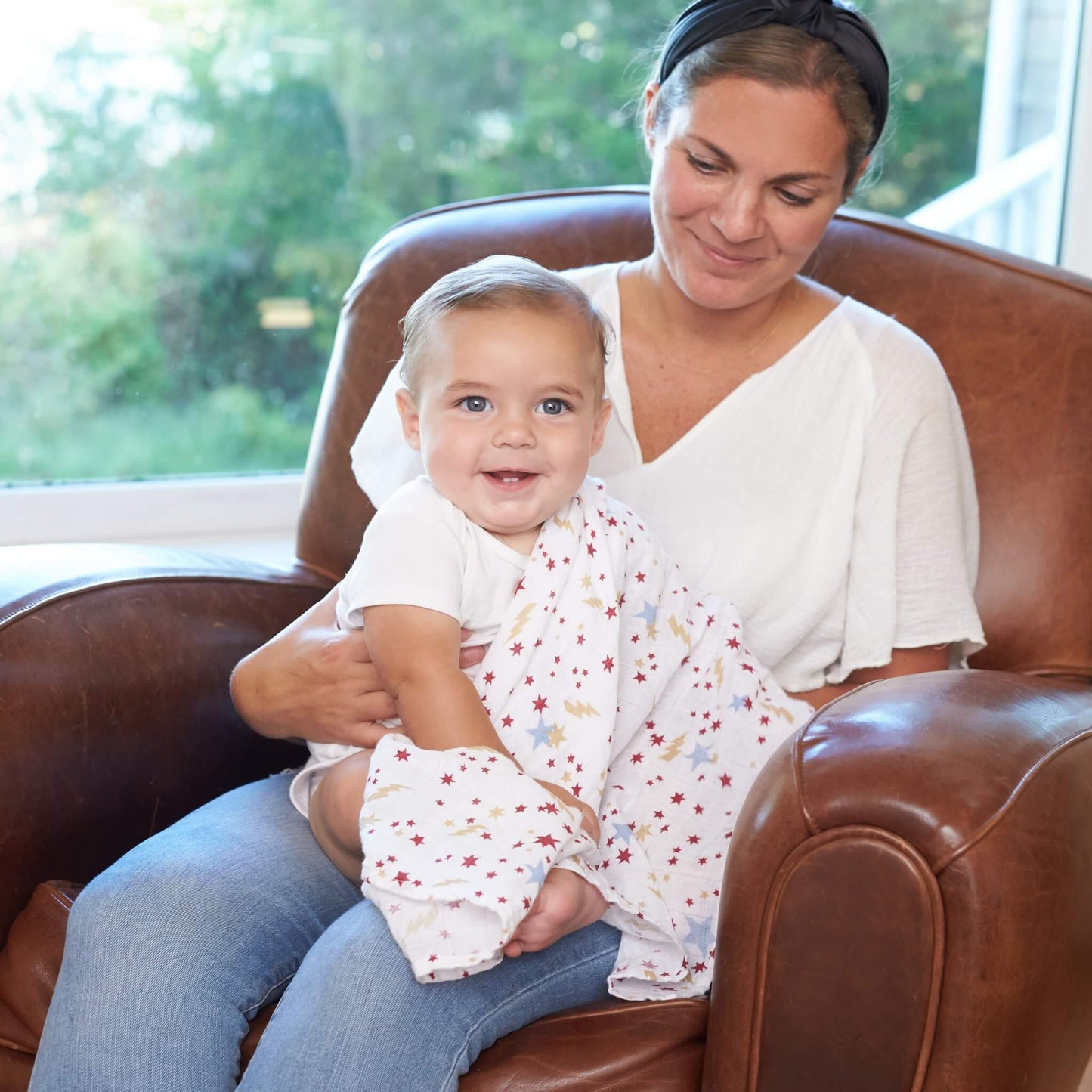 Mother holding smiling baby wrapped in the aden + anais Harry Potter™ muslin swaddle, seated together on a leather chair.