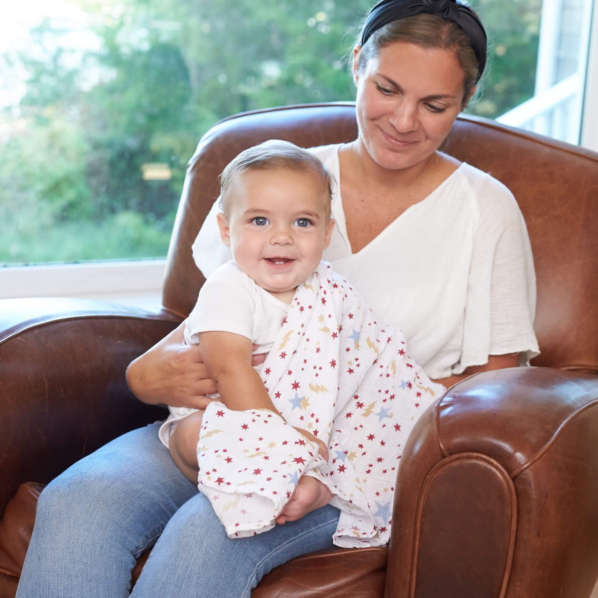 Mother holding smiling baby wrapped in the aden + anais Harry Potter™ muslin swaddle, seated together on a leather chair.