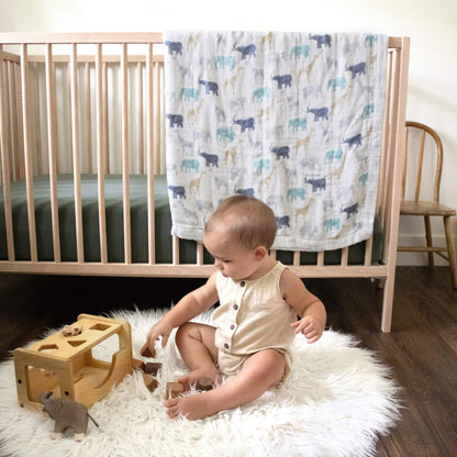 Baby sitting on a rug beside a cot, with the aden + anais Expedition Dream Blanket draped behind. A soft, cosy layer for play or sleep.