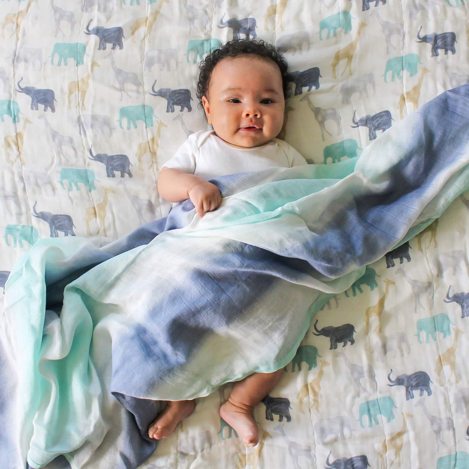 Smiling baby lying on the aden + anais Expedition Dream Blanket. Colourful elephant and giraffe print on breathable bamboo muslin.