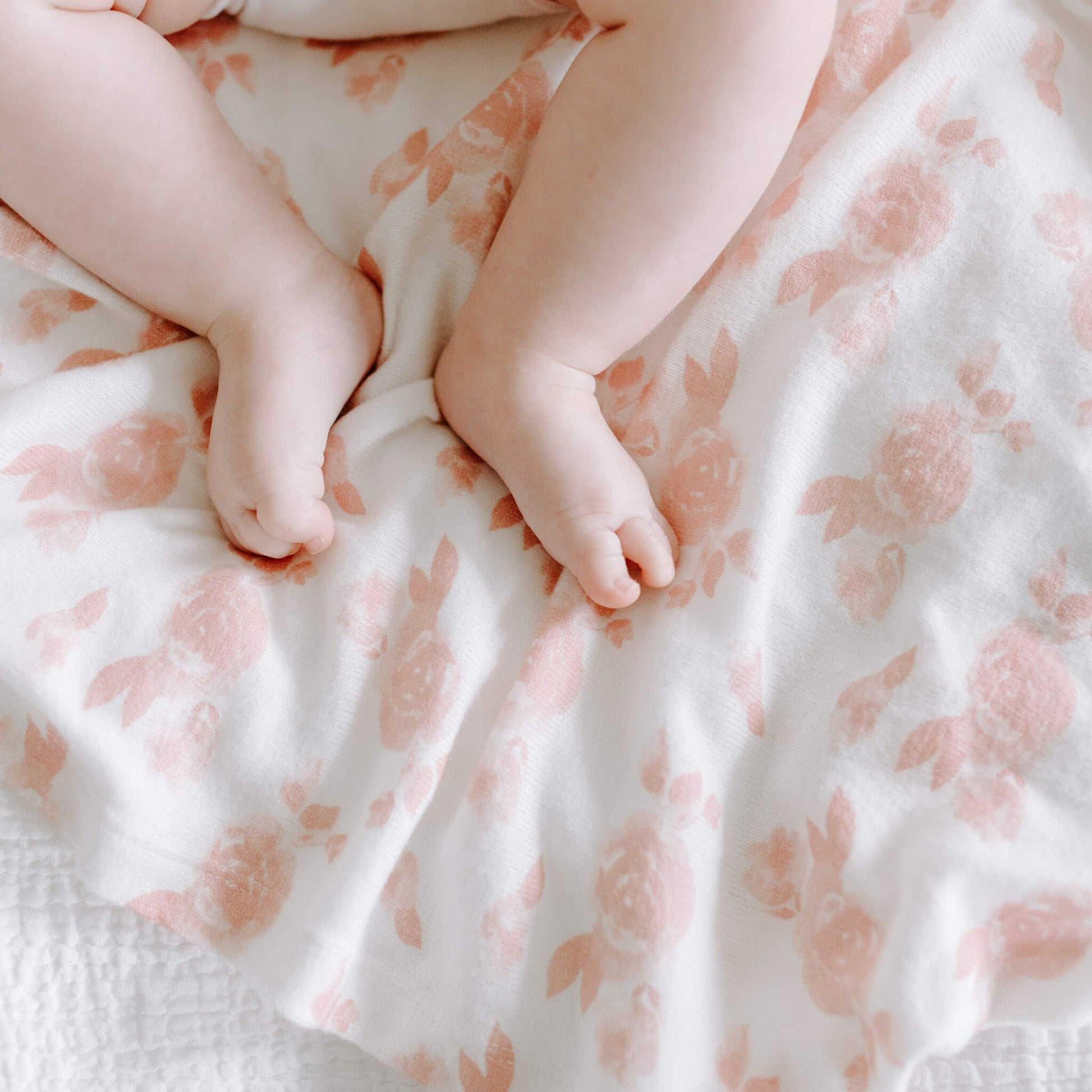 Close-up of baby’s feet resting on the aden + anais Rosettes swaddle. Soft jersey knit with gentle pink floral pattern.