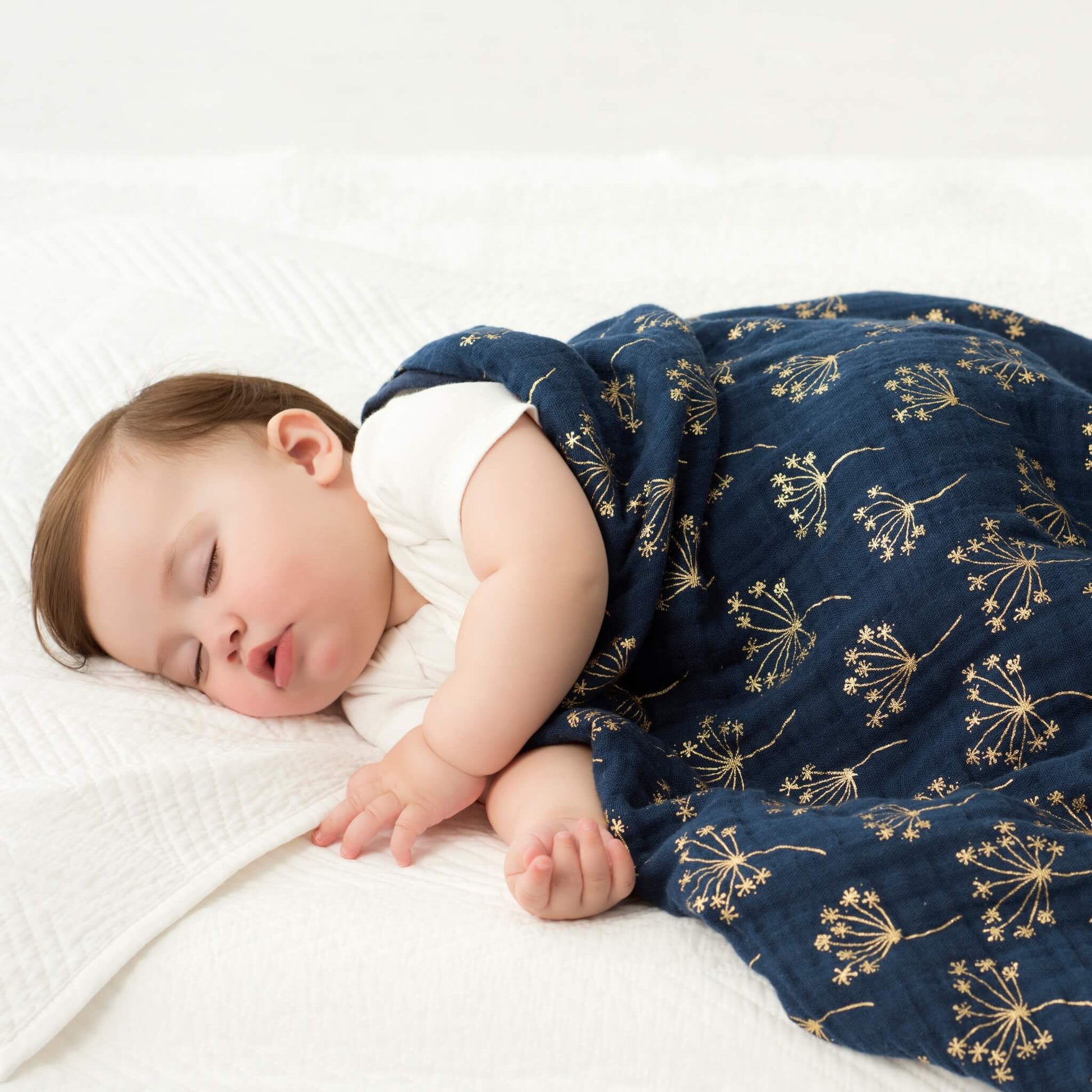 Sleeping baby snuggled under the aden + anais Metallic Gold Deco Dream Blanket, featuring a dark blue and gold floral print.