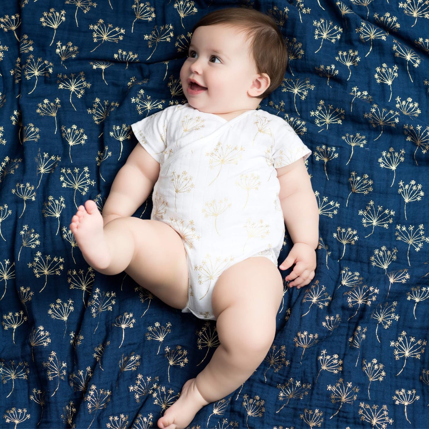 Baby smiling while lying on the aden + anais Metallic Gold Deco Dream Blanket, surrounded by gold floral patterns on a navy muslin base.