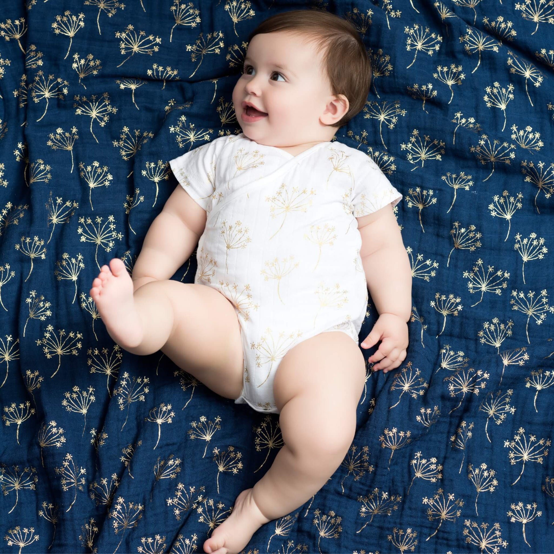 Baby smiling while lying on the aden + anais Metallic Gold Deco Dream Blanket, surrounded by gold floral patterns on a navy muslin base.