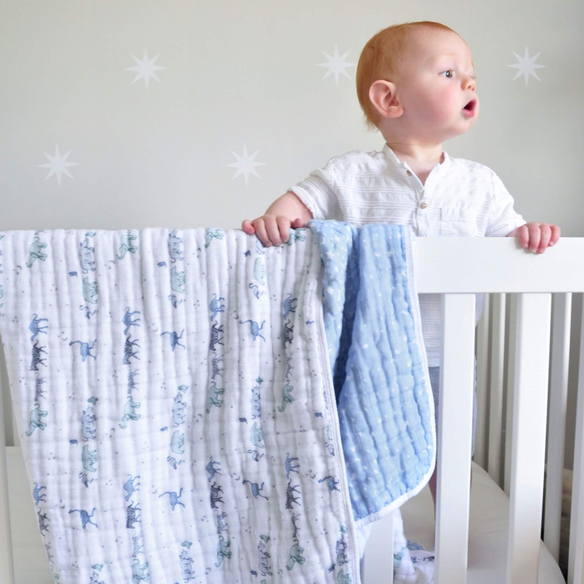 Baby standing at a white cot with the aden + anais Rising Star Dream Blanket draped over the side, showing soft cotton muslin in safari and star prints.