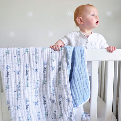 Baby standing at a white cot with the aden + anais Rising Star Dream Blanket draped over the side, showing soft cotton muslin in safari and star prints.