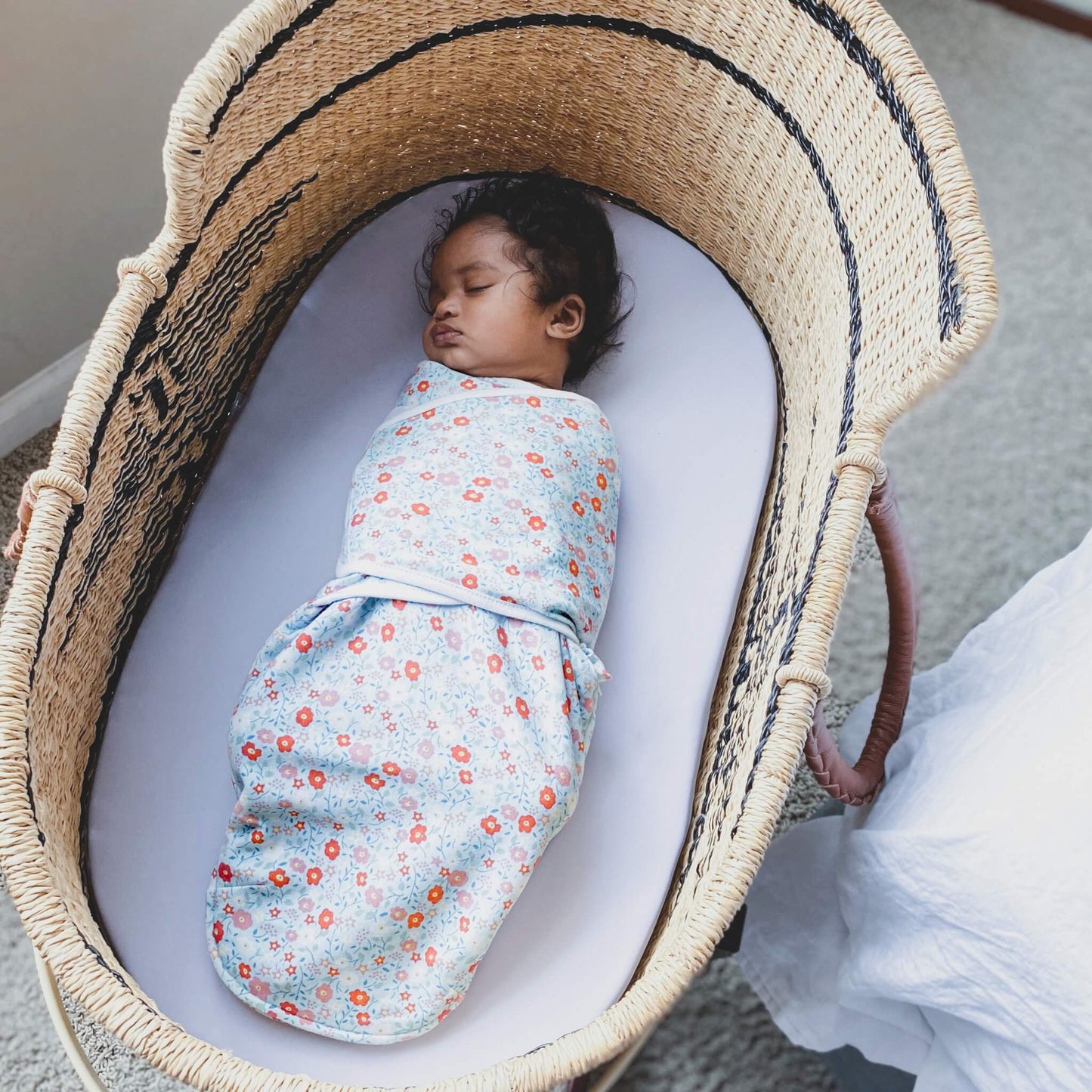 Baby sleeping peacefully in a woven Moses basket, wrapped in aden + anais floral print swaddle.