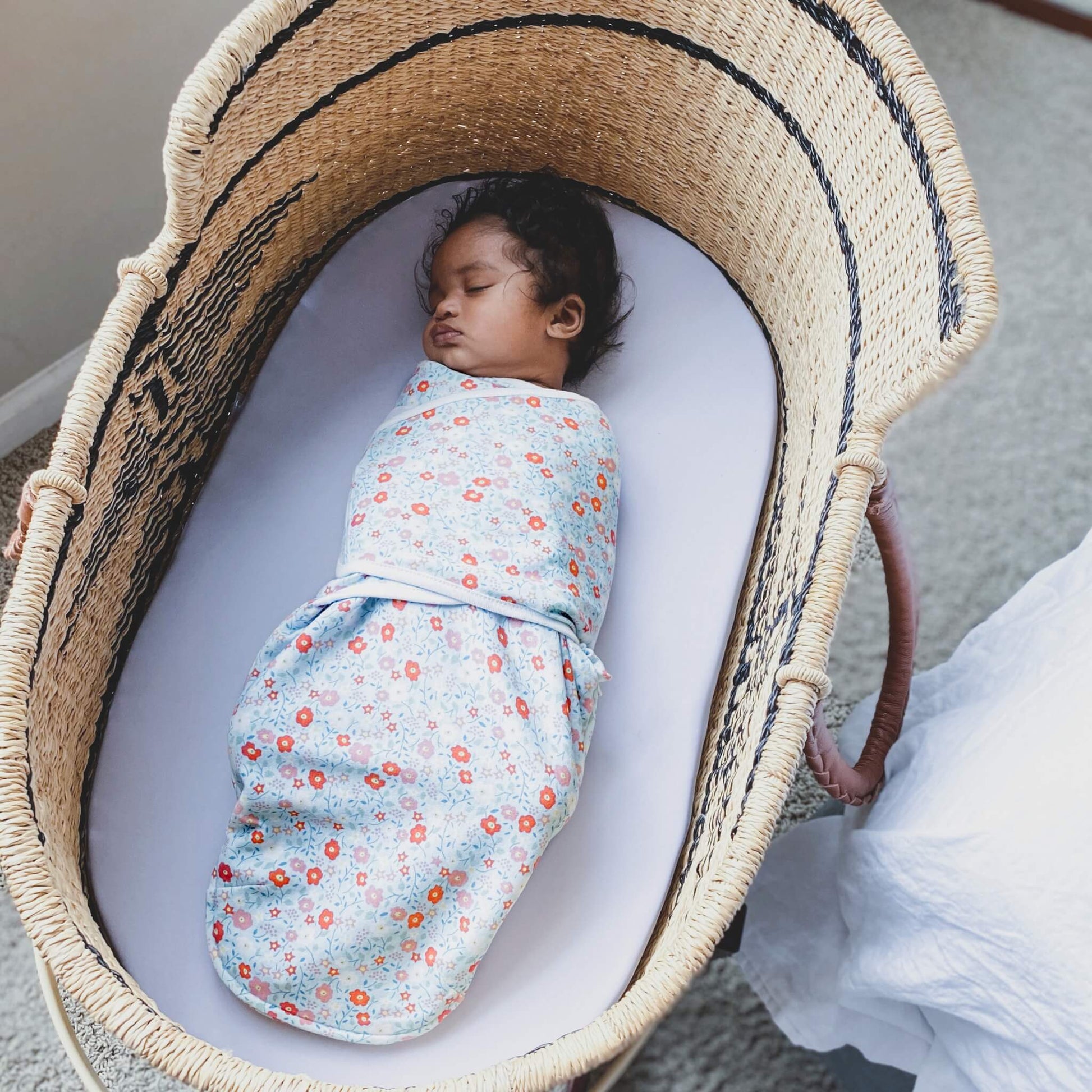Baby sleeping peacefully in a woven Moses basket, wrapped in aden + anais floral print swaddle.