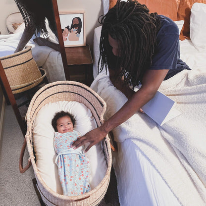 Parent gently touching baby swaddled in floral aden + anais wrap, lying happily in a Moses basket.