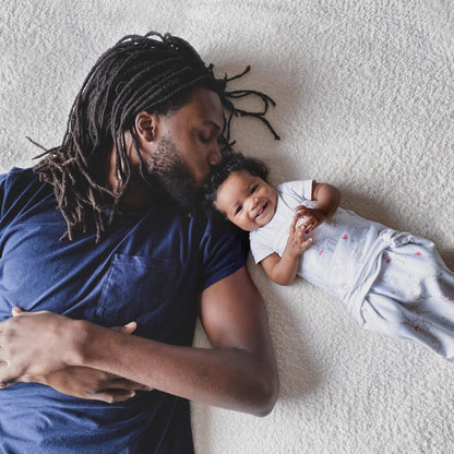 Father lying next to smiling baby wearing aden + anais woodland animal print wrap swaddle on a cosy bed.
